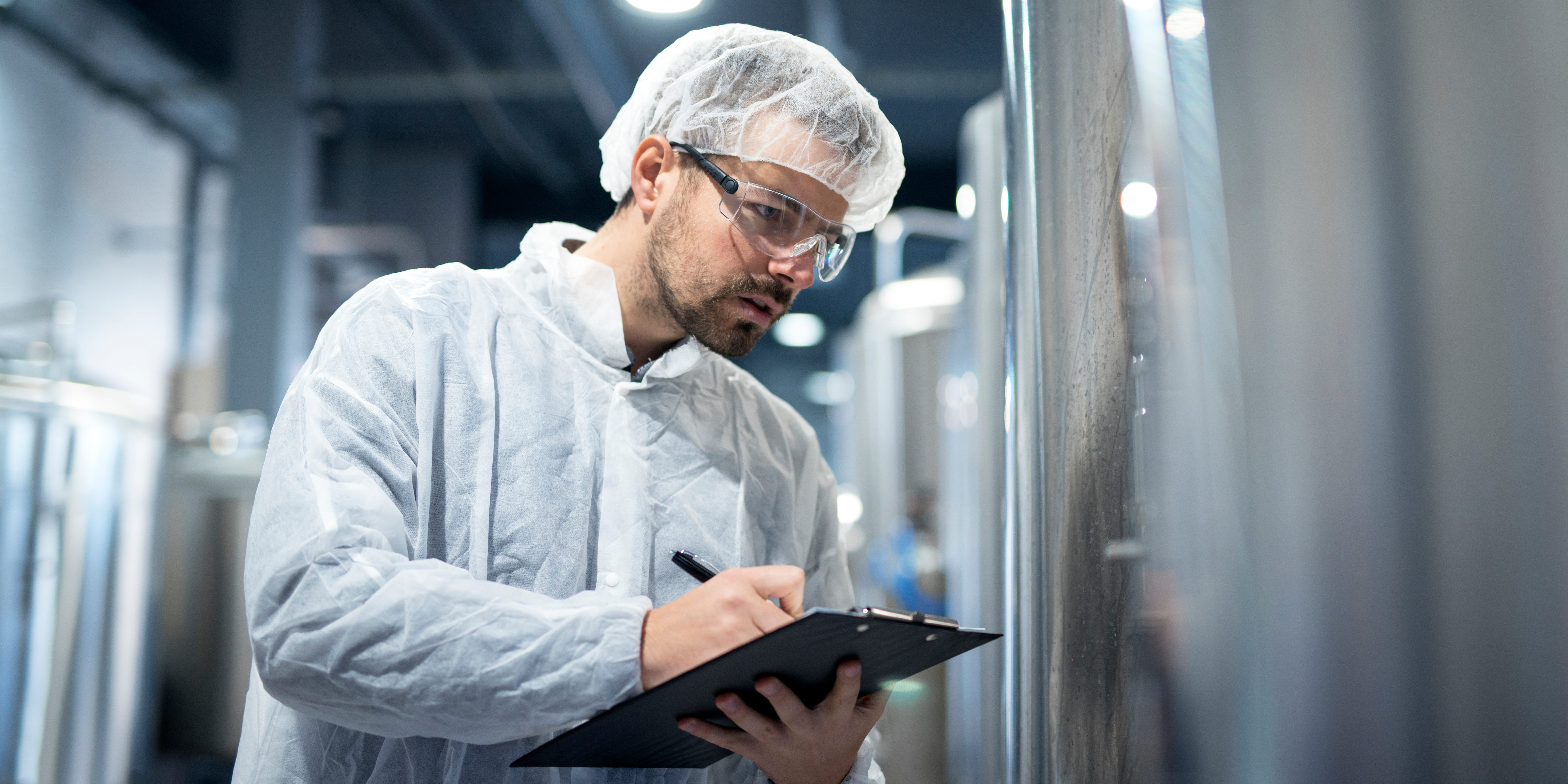 Technicien en tenue de protection inspectant une cuve industrielle et notant ses observations sur une tablette